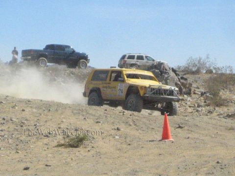 1990 Ford Bronco - yellow off-road race SUV grinds along a rocky desert trail with pickups and an SUV parked in the background.