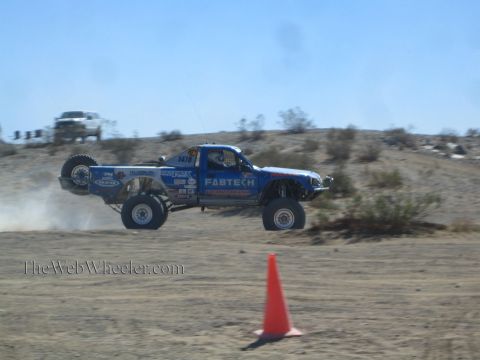 1990 Ford Bronco - blue Fabtech-sponsored off-road race truck launches off a desert jump, all four wheels in the air above a traffic cone.