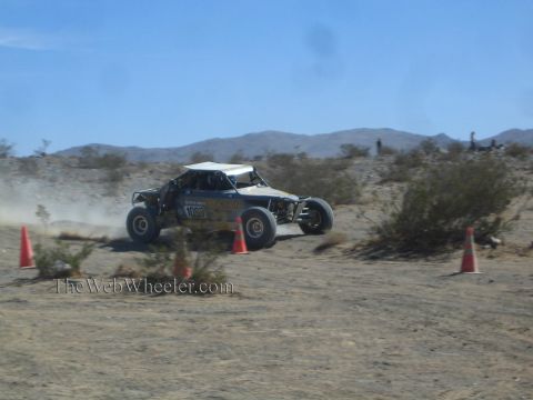 1990 Ford Bronco - open-cockpit single-seat off-road race buggy speeds across flat desert terrain between traffic cones.