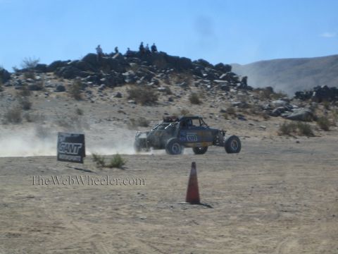 1990 Ford Bronco - tube-frame off-road race truck races a desert course past a sponsor banner as spectators sit on a rocky ridge.