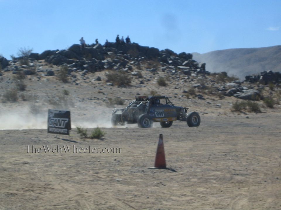Tube-frame off-road race truck on a desert course past a banner and cone