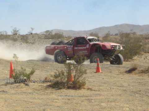 1990 Ford Bronco - red off-road race truck marked 1404 powers through a sandy desert course past orange traffic cones.