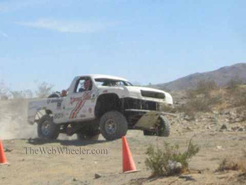 1990 Ford Bronco - white off-road race truck marked 801 corners hard through a desert course, spraying dirt near traffic cones.