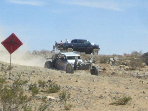 1990 Ford Bronco - open-frame off-road race buggy climbs a rocky desert grade as spectators watch from a pickup atop the ridge.