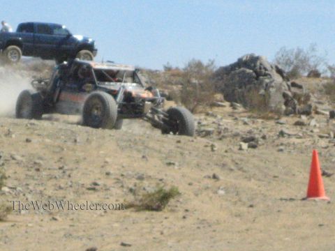 1990 Ford Bronco - orange and silver tube-frame off-road race buggy climbs a rocky desert grade past a cone with a pickup parked above.