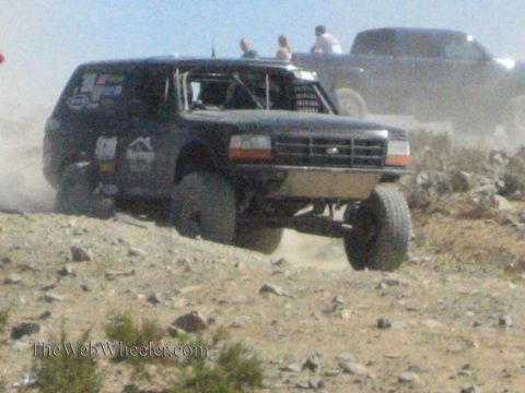 1990 Ford Bronco - dark Ford-badged off-road race truck flexes its suspension climbing a rocky desert grade as spectators watch.