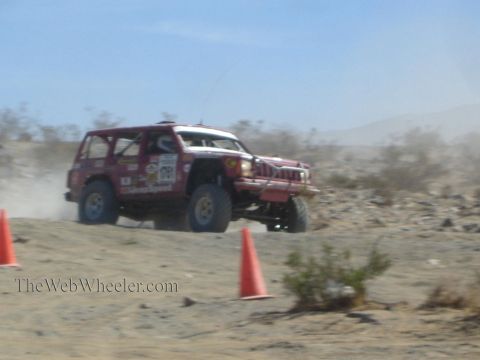 1990 Ford Bronco - red and white off-road race SUV crests a desert rise with one wheel lifted, racing between orange traffic cones.