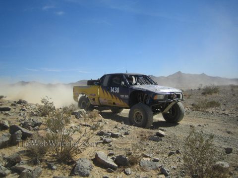 1990 Ford Bronco - blue and yellow off-road race truck marked 1438 runs a rocky desert trail under a clear blue sky.