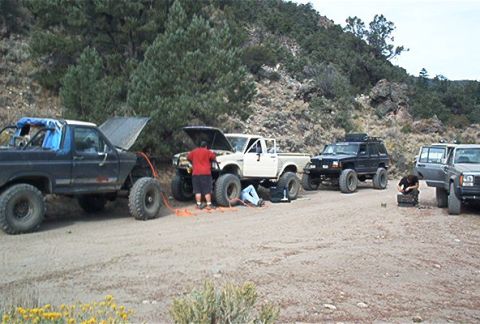 1986 Ford Bronco - Multiple off-road vehicles, including a 1986 Ford Bronco, are stopped on a dirt road with their hoods open as people work on them.
