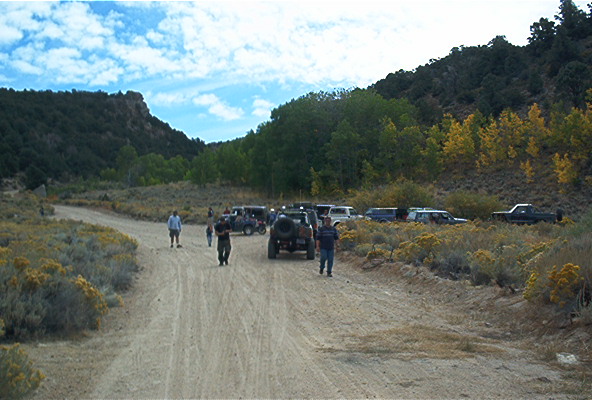 Several off-road vehicles parked and driving on a dirt road in a hilly, wooded area