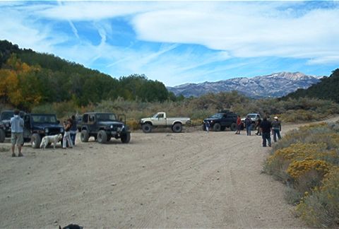1986 Ford Bronco - A group of off-road vehicles and people gather on a dirt road, with mountains visible in the distance under a cloudy sky.