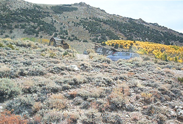 Blue SUV parked on a dry, brush-covered hillside near abandoned structures and yellow trees