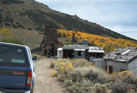1986 Ford Bronco - A blue pickup truck is parked on a dirt road in front of old, dilapidated buildings in a mountainous, wooded area.