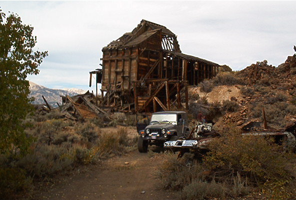 Dark gray Jeep Wrangler parked on dirt road near abandoned wooden building, mountains in background