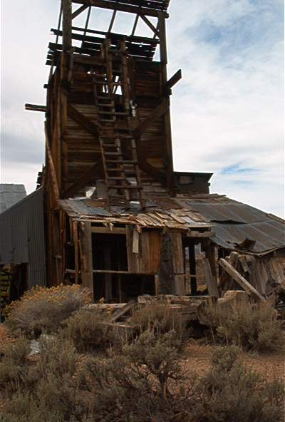 1986 Ford Bronco - An old, decaying wooden mine building stands against a cloudy sky, surrounded by dry brush.