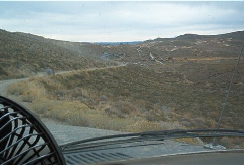 1986 Ford Bronco - A vehicle navigates a dusty, remote road through a barren, mountainous terrain under a cloudy sky.