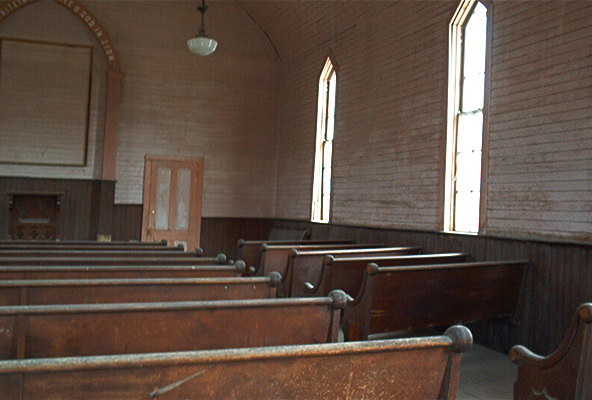 Interior of a weathered church with rows of wooden pews and arched windows