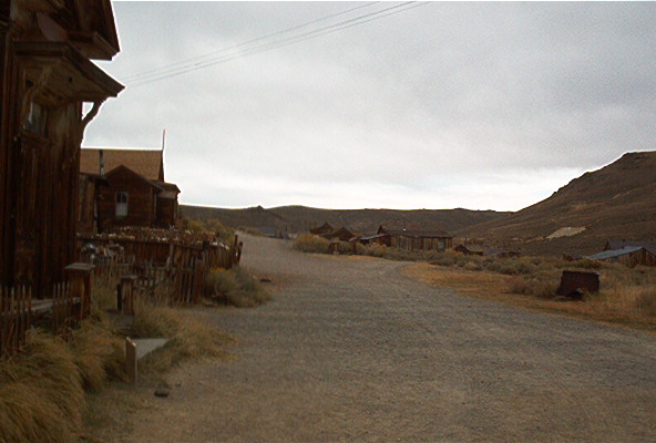Dirt road through a ghost town with wooden buildings and hills under a cloudy sky