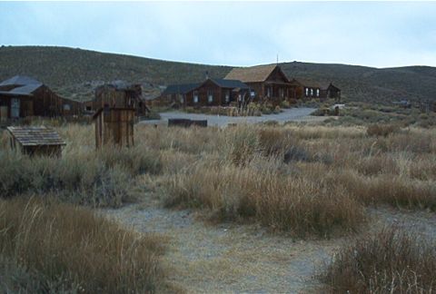1986 Ford Bronco - A desolate ghost town scene with weathered wooden structures and overgrown dry grass under a cloudy sky.