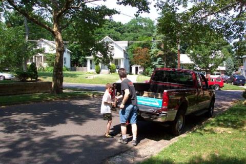 1979 Ford Bronco - Two people are loading items into the bed of a red pickup truck parked on a residential street.