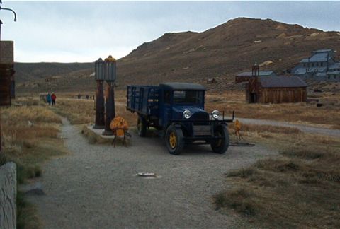 1986 Ford Bronco - A vintage blue flatbed truck sits parked next to antique gas pumps in a deserted town setting.