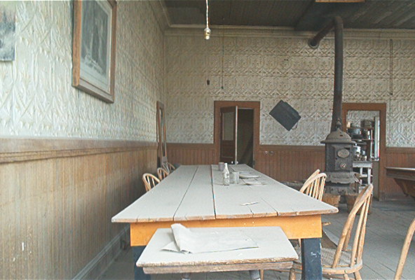 Interior of a rustic room with a long table and chairs, a wood stove, and framed pictures on the wall