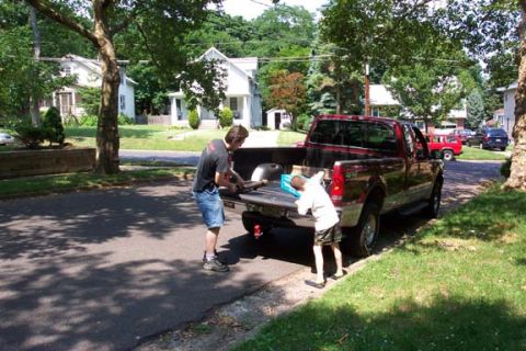 1979 Ford Bronco - Two individuals are seen loading items into the bed of a red pickup truck parked on a residential street.
