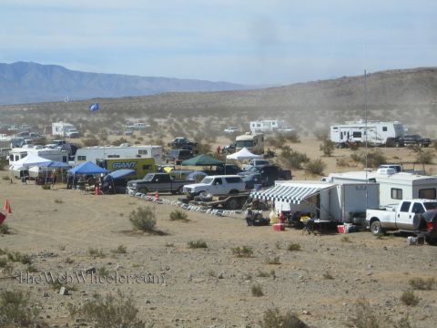 1990 Ford Bronco - RVs, trailers and pop-up canopies line a desert pit area at an off-road racing event under hazy skies.