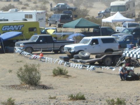 1990 Ford Bronco - Pickups and a white Ford Bronco on a trailer sit among RVs and tents in a desert off-road race pit area.