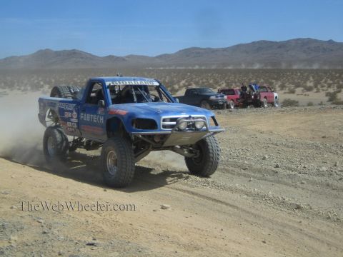 1990 Ford Bronco - blue Fabtech-liveried off-road race truck charges up a desert trail with other race vehicles trailing behind.