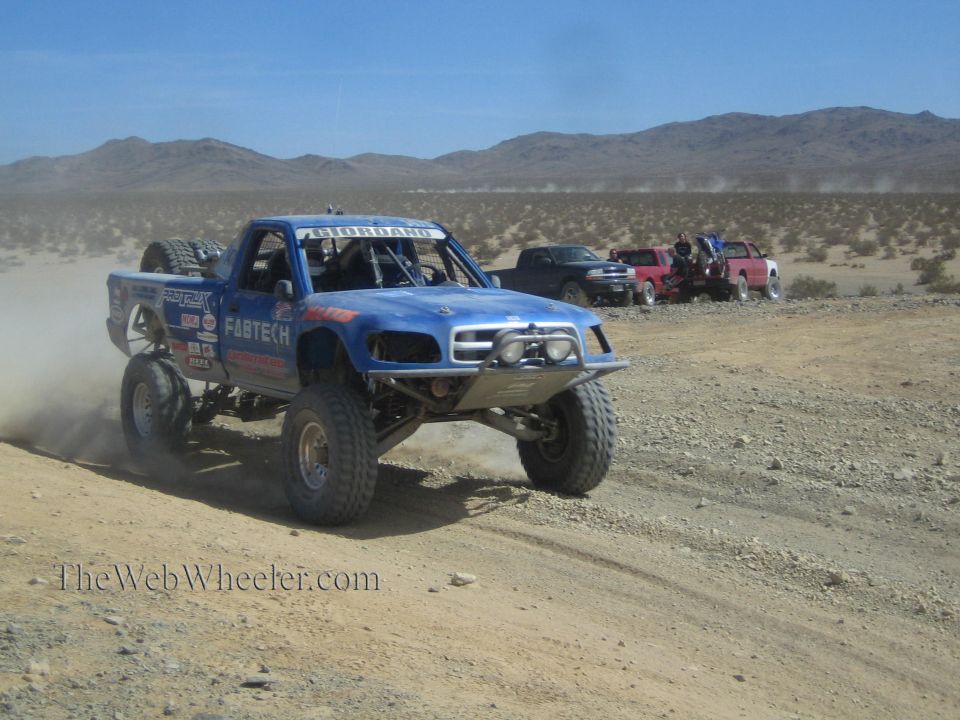 Blue Fabtech off-road race truck on a desert trail with other racers behind