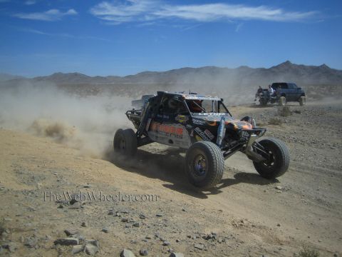 1990 Ford Bronco - open-frame off-road race buggy corners hard on a desert trail, throwing dirt as another racer follows behind.