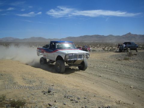 1990 Ford Bronco - vintage white and blue off-road race pickup runs a desert trail as chase trucks and racers gather behind.