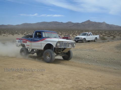 1990 Ford Bronco - white and red number 1487 off-road race truck speeds along a desert dirt track, trailing dust as a white pickup follows behind under distan…