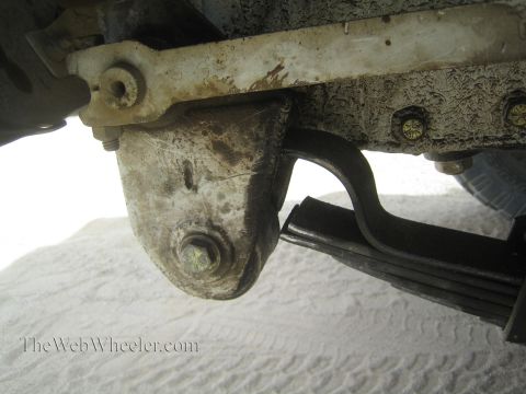 1990 Ford Bronco - grimy close-up of a leaf-spring shackle and bracket mount on a vehicle's undercarriage, with sand visible in the background.