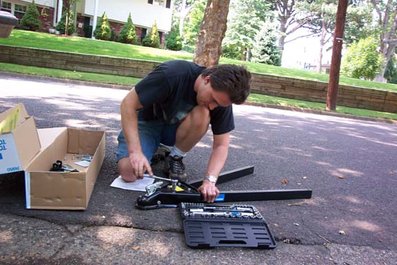 Man kneeling on asphalt, assembling a tow hitch with tools and parts from a box