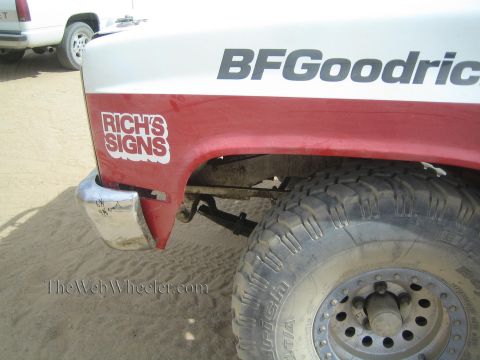 1990 Ford Bronco - close-up of a white and red off-road truck fender bearing BFGoodrich and Rich's Signs decals above a mud tire, parked on sand.