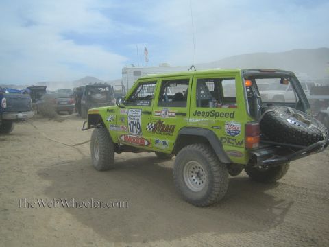 1990 Ford Bronco - lime green lifted Jeep Cherokee with JeepSpeed and sponsor decals, number 1719, sits at a dusty desert race staging area among trailers.