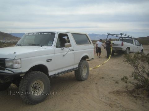 1990 Ford Bronco - white lifted Ford Bronco connected by a tow strap to a white pickup on a desert dirt road, with two people standing between the vehicles.