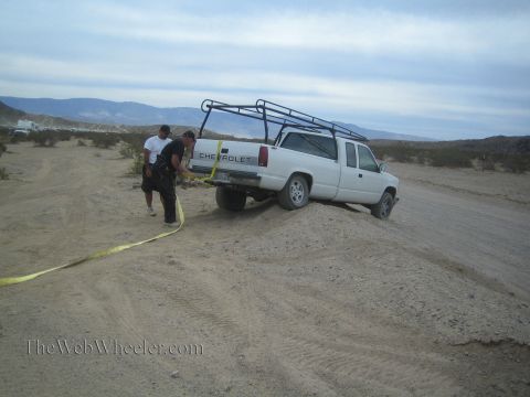 1990 Ford Bronco - white Chevrolet extended-cab pickup with a roof rack sits stuck in a sandy desert berm as two people work a yellow tow strap.