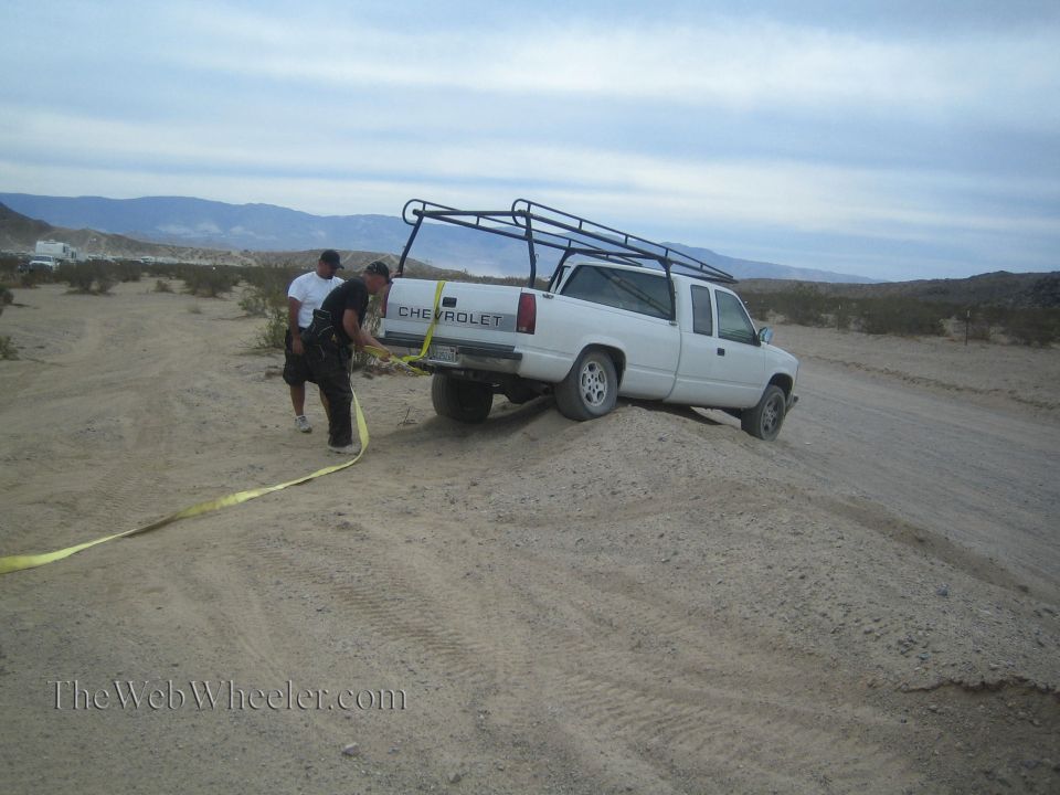 White Chevrolet pickup with roof rack stuck in desert sand being recovered