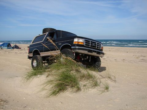 1996 Ford Bronco - A lifted 1996 Ford Bronco is parked on a sandy dune near the ocean with tents set up in the distance.