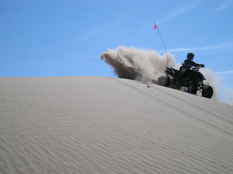1993 Ford Bronco - A rider on a quad bike navigates a sandy dune, creating a plume of dust against a clear blue sky.