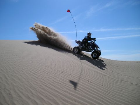 1993 Ford Bronco - A rider on a quad bike navigates a sandy dune, creating a plume of dust against a clear blue sky.