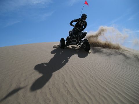 1993 Ford Bronco - A quad bike rider navigates a sandy dune, creating a cloud of dust against a clear blue sky.