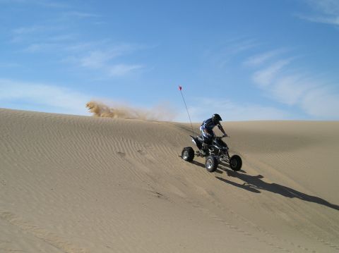 1993 Ford Bronco - A rider on a quad bike navigates a sandy slope, creating a dust trail against a bright blue sky.