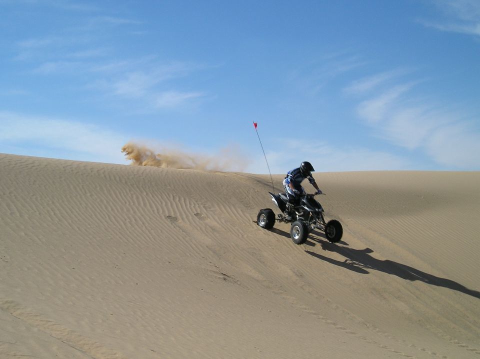 Rider on a black sport quad with sand tires cresting a dune, red whip flag flying