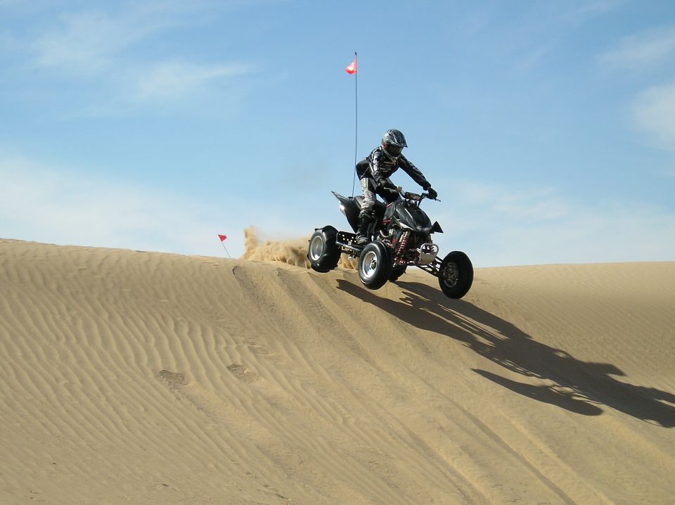 Rider jumping a black sport quad with safety whip flag off a sand dune crest
