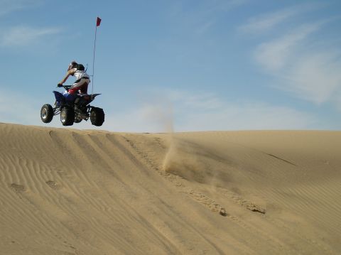 1993 Ford Bronco - An ATV rider catches air while cresting a sand dune under a blue sky.