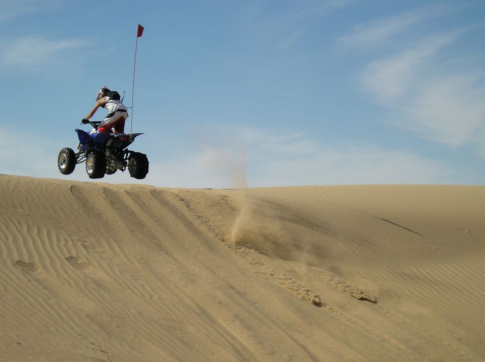 Blue sport quad ATV jumping off a sand dune crest with a red whip flag